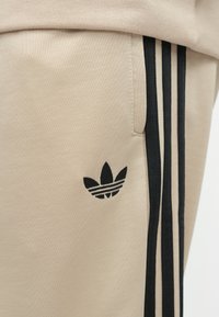 Μπεζ αθλητικό παντελόνι με μαύρες κάθετες ρίγες στο πλάι. Διαθέτει μαύρο λογότυπο Adidas στον μηρό, κατασκευασμένο από απαλό, υφασμένο ύφασμα.
