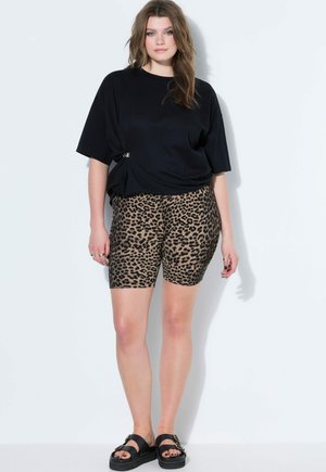 Vrouw die een zwart oversized T-shirt draagt, biker shorts met luipaardprint en zwarte platformsandalen, staand tegen een effen witte achtergrond.