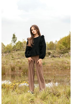 Zwarte, gestructureerde, langermige top met ruches, gecombineerd met losse, lichtbruine wijde broek. Buitenscène met groen in de achtergrond.