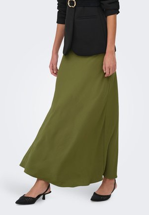 Femme portant une longue jupe vert olive, un blazer noir ceinturé et des talons slingback noirs pointus, debout devant un fond uni.
