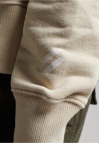 Beige mikina se žebrovanými manžetami a měkkou texturou, s bílým logem na rukávu. Tkanina vypadá silně a teple.