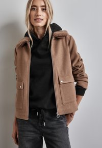 Chaqueta de forro polar marrón con un cuello texturizado, dos bolsillos frontales y cierre de cremallera. Llevada sobre una sudadera con capucha negra.