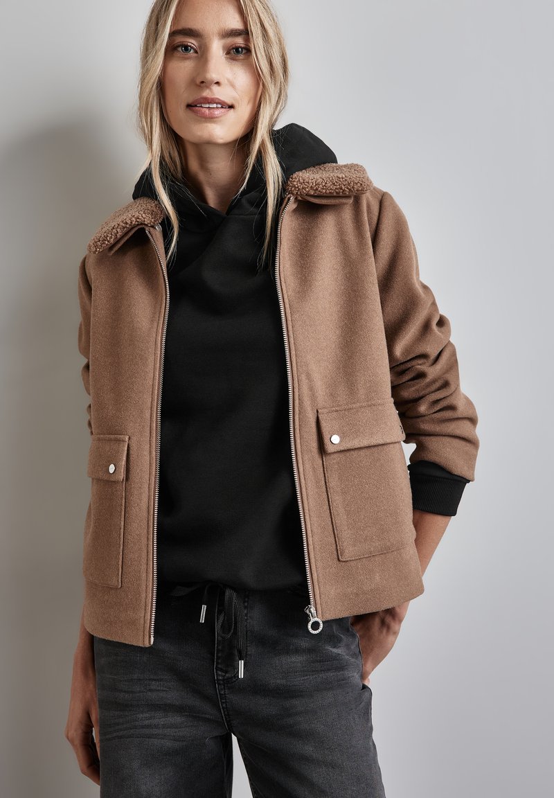 Chaqueta de forro polar marrón con un cuello texturizado, dos bolsillos frontales y cierre de cremallera. Llevada sobre una sudadera con capucha negra.