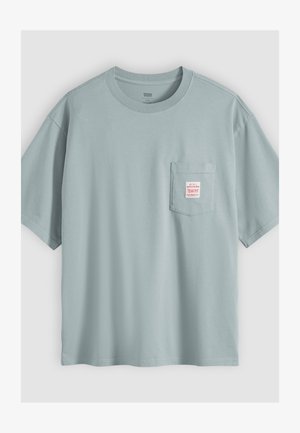 Vaaleansininen lyhythihainen puuvillainen t-paita, jossa on rintatasku ja pieni valkoinen etiketti punaisella tekstillä.