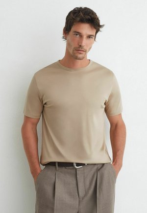 Man met kort donker haar, gekleed in een beige shirt met korte mouwen en grijze geplooide broek, staand met handen in de zakken tegen een witte muur.