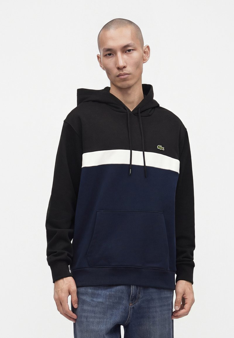 Homme portant un sweat à capuche noir avec des blocs de couleur blanc et bleu marine et un petit logo vert sur la poitrine, associé à un jean bleu, debout devant un fond blanc.