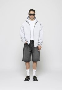 Šviesiai pilka džemperis su užtrauktuku, balti marškinėliai, tamsiai pilkos džinsinės šortai, balti kojinės, juodi loaferiai ir juodos akiniai nuo saulės. Tvarkingas, kasdienis dizainas.