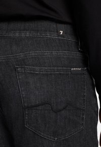 Des jeans en denim noir comportent une poche arrière avec un motif de couture ondulée, une étiquette de marque et une finition de texture lisse.