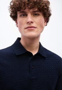 Νεαρός άνδρας με καστανά σγουρά μαλλιά που φοράει σκούρο μπλε πουκάμισο πόλο με υφή, κοιτάζοντας μπροστά σε λευκό απλό φόντο.