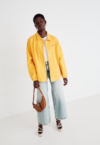 Gul oversized denimjakke med trykknapper, brukt over en hvit skjorte; sammen med lyseblå vide jeans og en brun håndveske.