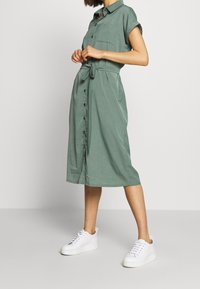 Donna in piedi che indossa un vestito midi verde con bottoni, fascia in vita e sneaker bianche con piattaforma, su uno sfondo neutro.