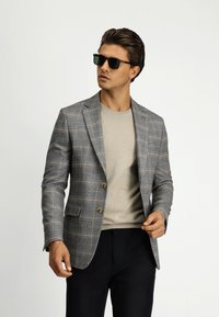 Grijze geruite blazer met twee knopen, over een beige trui, gecombineerd met zwarte broek. Het model draagt een donkere zonnebril.