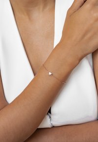Rose gold bracelet featuring a small heart charm, delicate chain design, and smooth finish, worn on a tan wrist against a white background.