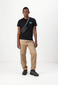 Tricou negru cu logo verde, pantaloni khaki și adidași negri. O mică geantă crossbody neagră este purtată pe umăr. Fundal neutru.