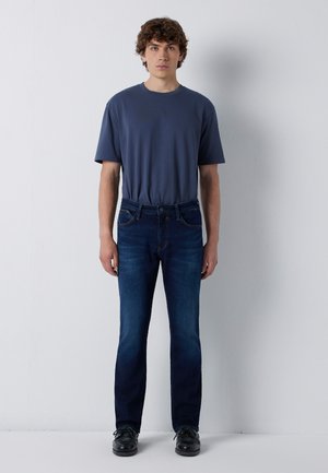 Young man with curly hair stands facing forward wearing a navy blue t-shirt, dark blue jeans, and black shoes against a plain white background.
