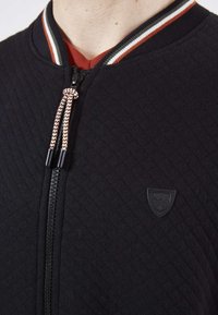 Veste bomber noire matelassée avec fermeture éclair, col rayé rouge, blanc et vert, et un petit écusson logo noir sur la poitrine.