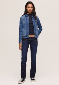 Jovem mulher vestindo uma jaqueta de ganga azul, jeans azul escuro, top cropped preto e ténis preto e branco, de pé contra um fundo simples.