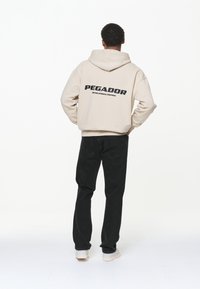 Sudadera con capucha beige con texto negro "PEGADOR" en la espalda, combinada con pantalones de mezclilla negros y zapatillas blancas. El material parece suave y con textura.
