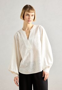 Blusa di cotone bianca con motivi a occhio ricamati, scollo a v, maniche voluminose e dettagli testurizzati, abbinata a pantaloni neri.