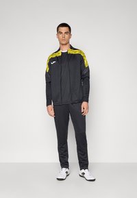 Zwarte trainingspak met gele accenten; het ritsjack heeft ribgebreide manchetten en een logo, gecombineerd met slim-fit broek en witte sneakers.