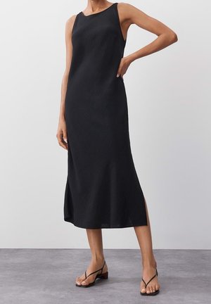 Kvinde iført en ærmeløs sort midi-kjole med sidelommer og sorte åbne hæle, stående mod en ensfarvet grå baggrund.