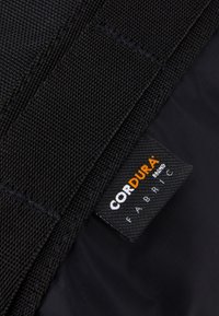 Svart, tätt vävt Cordura-tyg med sydda remmar och en liten svart etikett som visar "CORDURA fabric" i vit och orange text.