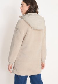 Beige fleece-takki, jossa on vaalea beige irrotettava huppu; sisältää ribattut hihansuut ja logon hihassa. Pehmeä tekstuuri, polven mittainen malli.