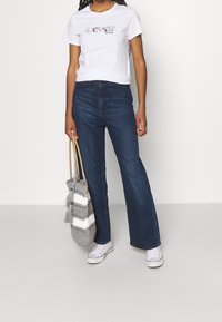 Camiseta blanca de manga corta con un diseño de "AMOR", combinada con unos jeans acampanados de color azul oscuro. Sosteniendo un bolso tejido de rayas con textura. Zapatillas blancas.