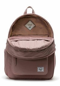 Sac à dos en rose poussiéreux avec un design à deux compartiments, fermeture éclair, poche frontale et étiquette en tissu. Texture lisse et forme arrondie.