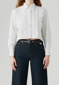 Chemise blanche courte avec un devant à boutons, des panneaux verticaux texturés et un col pointu, associée à un jean bleu foncé taille haute.