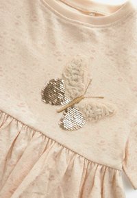 Robe rose pâle à manches longues ornée d'une application papillon pailletée avec des ailes texturées, et un ourlet de jupe plissé à volants. Tissu doux.