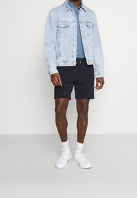 Ljust blå denimjacka, marinblå shorts, vita sneakers och vita strumpor. Jackan har framfickor och knappstängning. Avslappnad klädsel.