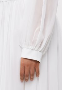 Weißes Kleid mit einer transparenten, puffartigen Ärmeln; glatter Stoff, Manschetten-Detail in reinem Weiß; Nahaufnahme einer Hand, die auf dem Stoff ruht.