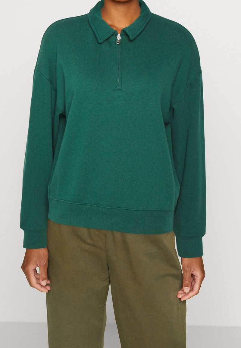 Maglione verde con collo a mezza zip, texture morbida, vestibilità rilassata e polsini a coste, abbinato a pantaloni color khaki.
