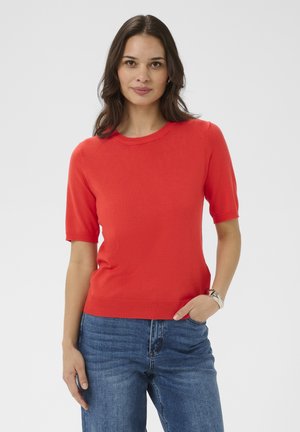 Femme aux cheveux noirs portant un pull rouge à manches courtes et un jean bleu, debout avec une main dans la poche devant un fond clair uni.