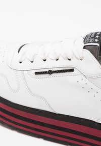 Sneaker bianco in pelle con dettagli perforati, chiusura con lacci e una suola spessa con strisce nere e rosse. Etichetta del logo sul lato.