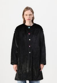 Črna umetna krznena plašč s krožnim izrezom, ki ima spredaj šest pisanih gumbov, podobnih dragim kamnom, in dolge rokave. Mehka tekstura.