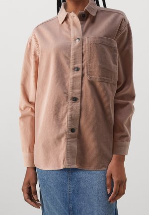 Buttoned long-sleeve pink corduroy shirt with chest pocket worn with blue denim skirt, model’s hands relaxed at sides.