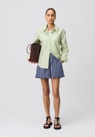 Camicia a righe verdi con bottoni, t-shirt bianca, pantaloni corti a righe blu navy, porta una borsa cilindrica marrone e indossa sandali neri.