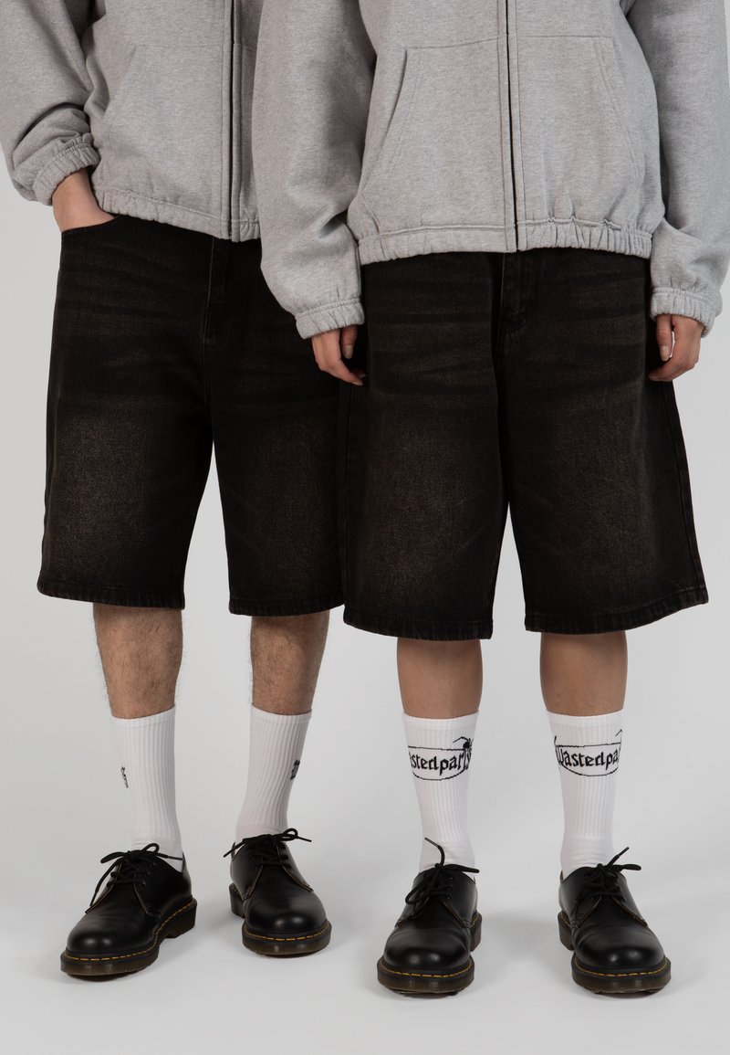 Shorts en denim noir avec une coupe décontractée, hoodie zippé gris, chaussures noires à lacets et chaussettes blanches avec texte. Ensemble simple et décontracté.