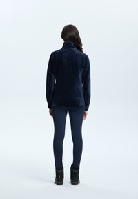 Marineblauwe fleecejack met hoge kraag en visgraatpatroon, gecombineerd met marineblauwe leggings en zwarte boots. Soepele textuur, aansluitende silhouet.