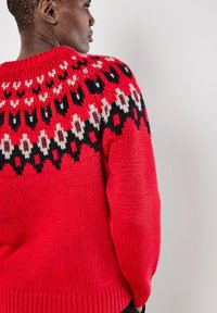 Roter Strickpullover mit einem gemusterten Halsabschluß in Schwarz, Weiß und Beige. Die Textur ist grob und hat einen gerippten Saum.