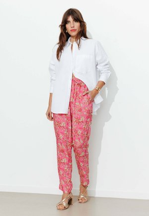 Femme debout contre un mur blanc, portant une chemise blanche à boutons, un pantalon rose à fleurs, un bracelet en or et des talons ouverts dorés.
