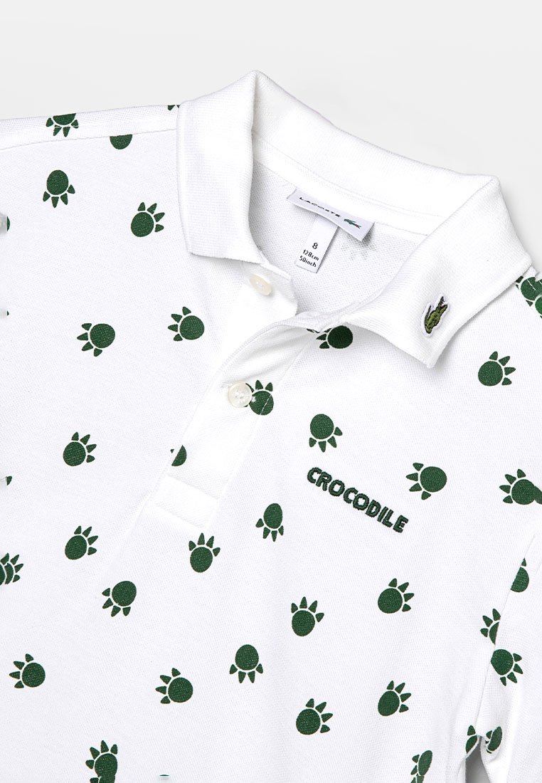Biała koszulka polo z powtarzającym się zielonym wzorem odcisku stopy żółwia. Wyszywane napisy "CROCODILE" oraz logo przy kołnierzyku.
