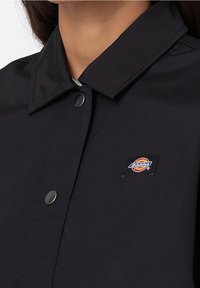 Camisa negra de botones con cuello puntiagudo, que presenta botones metálicos y un pequeño parche con el logo de Dickies en la parte delantera. Textura suave.
