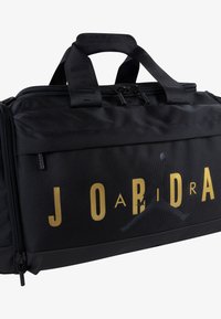 Bolsa de deporte negra hecha de tela duradera; presenta letras y un logo dorados, asas resistentes y un cierre de cremallera para un almacenamiento seguro.