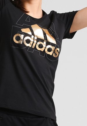 Persoon draagt een zwart Adidas t-shirt met een groot goud logo, steekt één arm omhoog tegen een effen lichtgrijze achtergrond.