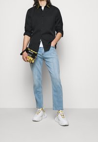 Camisa negra de botones con cuello estampado, jeans azules claros y zapatillas blancas con acentos amarillos, llevando un bolso negro y dorado.