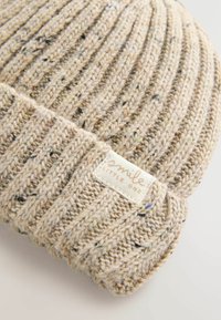 Dziergana czapka beanie w jasnym beżu z teksturowanym wzorem ściegu. Posiada małą metkę z napisem "uśmiech" i "maluch". Miękki materiał.