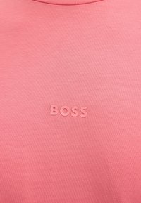 Ροζ ύφασμα με ανυψωμένα γράμματα που σχηματίζουν τη λέξη «BOSS», τοποθετημένα στο κέντρο κοντά στο πάνω μέρος, υποδηλώνοντας ένα επώνυμο ένδυμα ή λεπτομέρεια ρούχου.